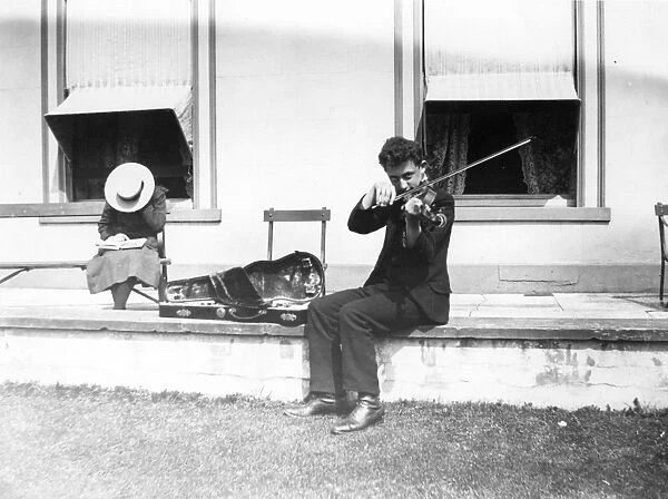 Violinist. circa 1908: A youth playing the violin, while sitting on a wall outsde a house