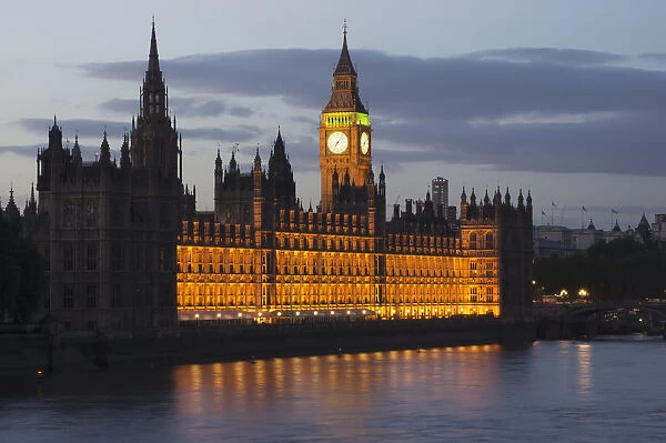 A Building And Clock Tower Along The Waters Edge Illuminated At Dusk; London, England