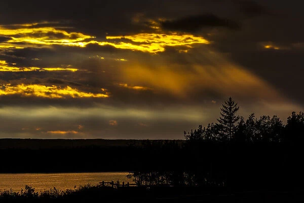 Dramatic colourful sky with sun beams coming down from the sky reflecting on a lake with trees silhouetted in the foreground; Calgary, Alberta, Canada