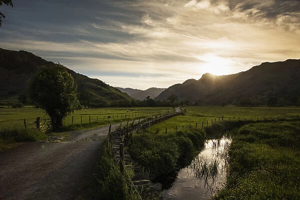 A Gravel Road Between Fields As The Sun Sets Behind The Mountains; Lake District, Cumbria, England