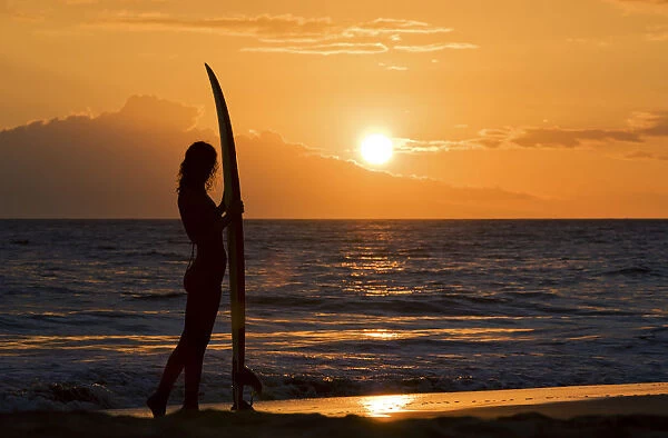 Hawaii, Female Surfer On Beach Silhouetted Against Orange Sunset Over Ocean