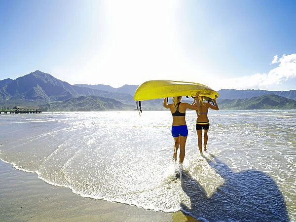 Hawaii, Kauai, Hanalei Bay, Two Active Young Woman Carrying A Kayak Into The Ocean
