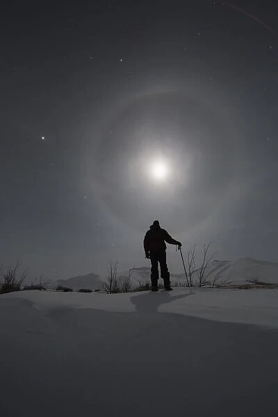 Man Snowshoeing Along The Dempster Highway Under A Moon Glowing With Rings; Yukon, Canada