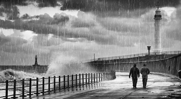 Two People Walking Along The Waters Edge In The Rain With Waves Crashing Into The Shore And A Lighthouse In The Background; Sunderland, Tyne And Wear, England