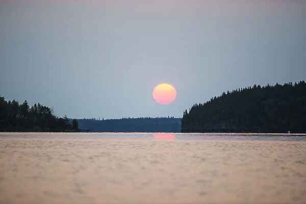 A Red Sun Setting Over The Forest With A Lake Reflecting The Pink Glow; Manitoba, Canada