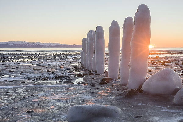 Rocks And Pilings Covered With Snow And Ice Along The Shore At The Waters Edge, With A View Of The Coastline And Mountains In The Distance; Alaska, United States Of America