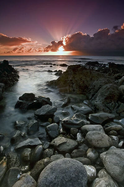 Sun Rays Shine Above The Clouds As The Sun Sets Over The Ocean With Rocks Along The Coast In The Foreground; Hawaii, United States Of America