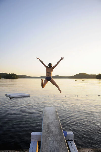 A Swimmer Jumps Off A Diving Board As The Sun Sets Over A Lake In Muskoka; Huntsville, Ontario, Canada