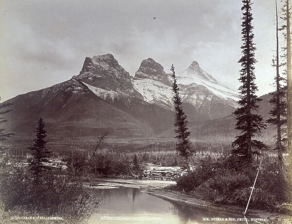 The three peaks known as The three sisters in the Rocky Mountains in Canada. In the foreground a river runs through the woods in the valley below