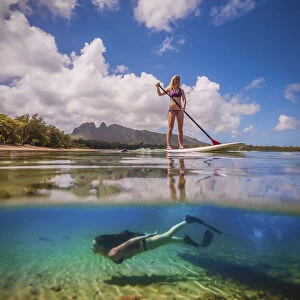 An athletic woman rides a stand-up paddle-board as another woman free-dives below her