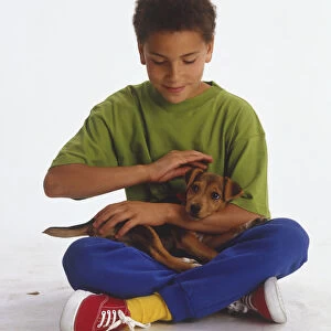 Boy wearing blue trousers and green t-shirt, sitting cross-legged on floor, cuddling and stroking small brown and black puppy