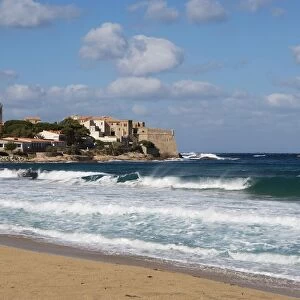 Corsica, Algajola, waves on rough seas with beach Citadelle on coastline in distance with blue sky and white clouds above