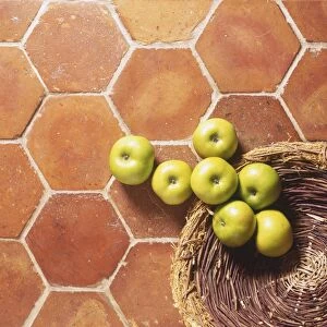 Green apples tumbling out of wicker basket onto tiled kitchen floor, close up, view from above