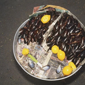 Overhead view of Black Sea Mussels arranged on a round tray covered with newspaper with lemons and a knife, a Turkish dish