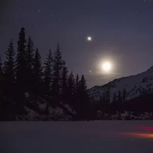 Nighttime View Of A Man Standing Next To A Lit Tent On A Snow Covered Trail Lake With The Moon, Stars, And Planets Overhead, Moose Pass, Kenai Peninsula, Southcentral Alaska