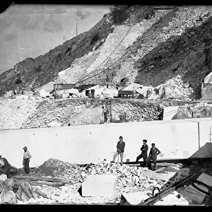 Three men observe the work of several excavators in the Apuan Alps. Behind them is an enormous block of marble already squared off and ready to be transported to the valley