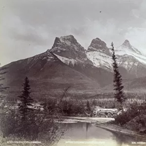 The three peaks known as The three sisters in the Rocky Mountains in Canada. In the foreground a river runs through the woods in the valley below